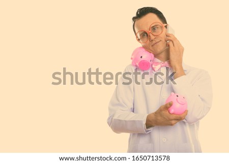 Studio shot of young crazy man doctor holding two piggy banks while talking on mobile phone