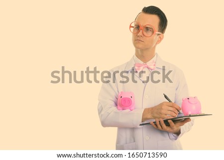 Studio shot of young crazy man doctor writing on clipboard while thinking with two piggy banks