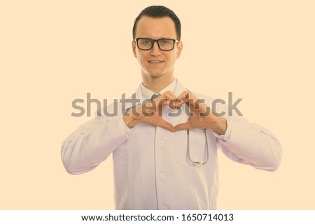 Studio shot of young happy man doctor smiling while giving hand heart gesture