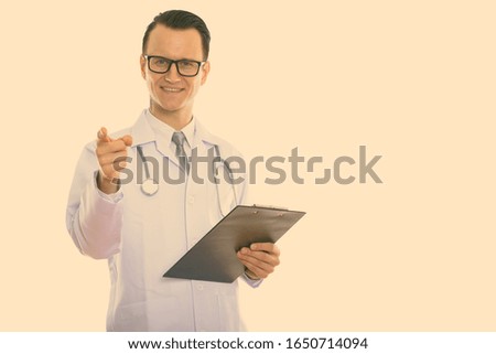 Studio shot of young happy man doctor smiling while holding clipboard and pointing finger at camera