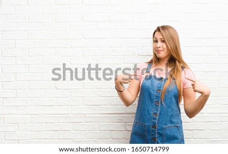 young blonde woman looking proud, positive and casual pointing to chest with both hands against brick wall