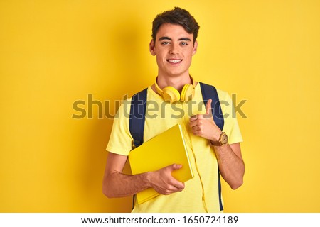 Teenager boy wearing headphones and backpack reading a book over isolated background happy with big smile doing ok sign, thumb up with fingers, excellent sign