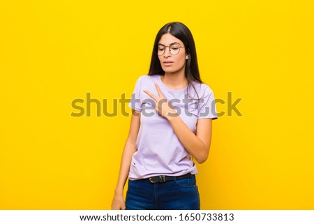 young pretty latin woman feeling happy, positive and successful, with hand making v shape over chest, showing victory or peace against flat wall