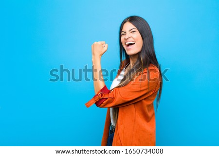 young pretty latin woman feeling happy, satisfied and powerful, flexing fit and muscular biceps, looking strong after the gym against flat wall Royalty-Free Stock Photo #1650734008