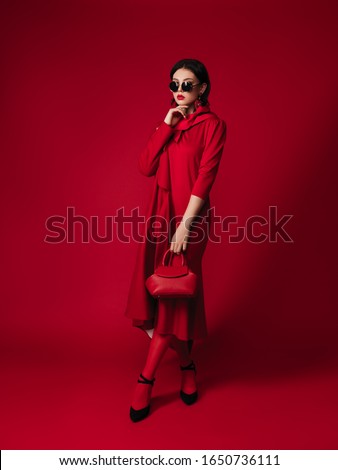 total red look style fashion studio portrait of a gorgeous young brunette woman, coat on one shoulder, dress, bag, round sunglasses, head scarf, makeup, brooch with feathers, red lips, high heels