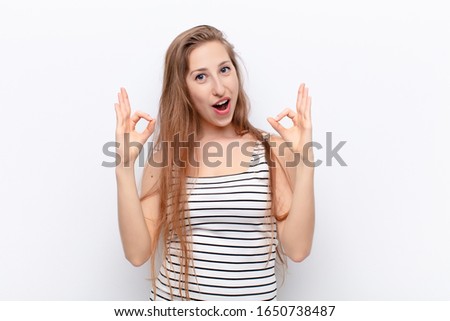 yound blonde woman feeling shocked, amazed and surprised, showing approval making okay sign with both hands against white wall