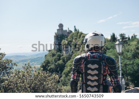 Back view. Man dressed in a motorcycle outfit and white helmet. body protection turtle. Fortress on top of the mountain background. Copy space. Motorbike travel. Pass of the witches San Marino