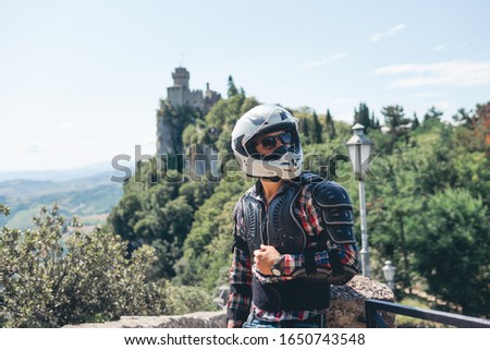 Man dressed in a motorcycle outfit and white helmet and sunglasses. body protection turtle. Fortress on background. Mountains. Stylish. copy space. Pass of the witches San Marino, Italy