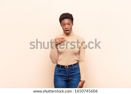 young pretty black woman feeling shocked, astonished and surprised, with hand on chest and open mouth, saying who, me?