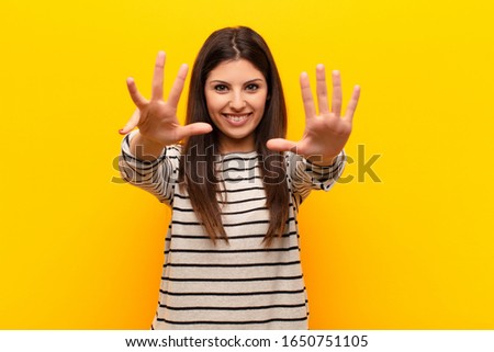 young pretty woman smiling and looking friendly, showing number nine or ninth with hand forward, counting down against yellow wall