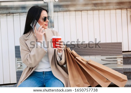 Stylish woman talking on the phone, drinking coffee and holding eco bags after doing shopping in the mall. Black Friday concept