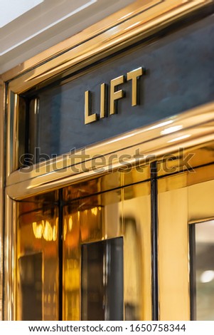 A stylish 'Lift' elevator sign with gold lettering on a dark background above a golden coloured elevator door