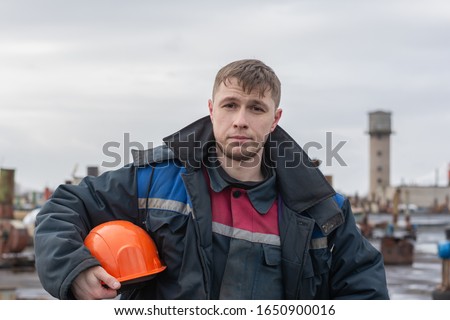 Worker builder on the roof of the factory on the background of electrical equipment on the street.  Holds an orange safety helmet under his arm.  Dressed in an old, shabby, dirty working uniform.  Royalty-Free Stock Photo #1650900016