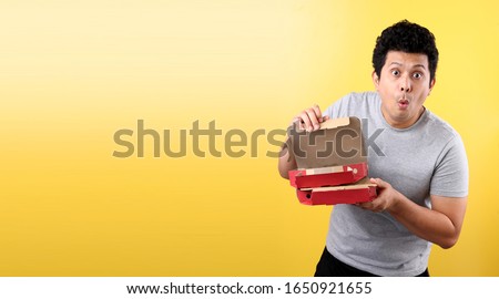 Young Asian man holding delivery box with Italian pizza scared in shock with a surprise face, on yellow background in studio With copy space.
