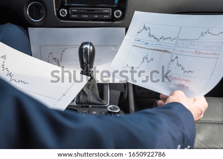 Businessman in the blue suit working in the car using laptop, graphics and charts printed on the paper 