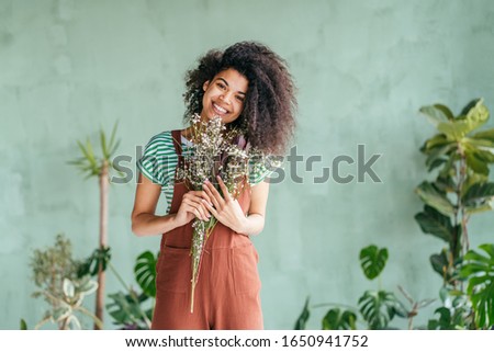Young woman cultivating home plants.Small business.Sensual mixed race female florist with flowers in hands against background of indoor plants. Life lover, zero waste, inspiration, summer mood concept Royalty-Free Stock Photo #1650941752