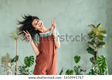 Eco friendly mixed race playful girl gardener fooling around shaking head with floating hair. African american woman holds hands with peace sign in brown overalls and strip t-shirt on green background