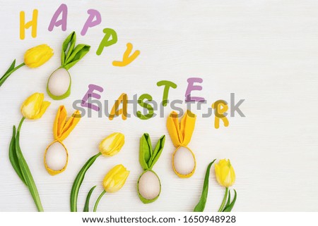 Easter composition with handmade colored eggs with ears from rabbit and word "Happy Easter" is made of colorful letters, fresh yellow flowers tulip.View from above, flat lay.