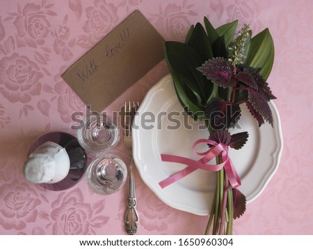Top view, on a pink tablecloth, a bouquet of green leaves and Coleus red flower on a white plate. Letter with the inscription with love and two wine glasses for wine. Place for text.