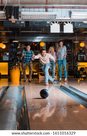 handsome young man throwing bowling ball on skittle alley near multicultural friends