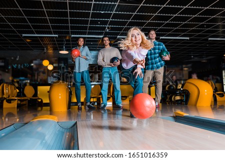 smiling blonde girl throwing bowling ball on skittle alley near multicultural friends