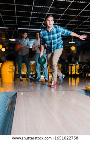 concentrated young man throwing bowling bowl on skittle alley near multicultural friends