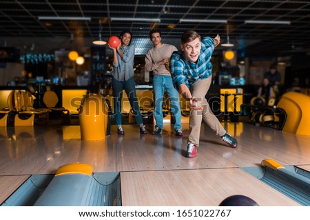 smiling young man throwing bowling bowl on skittle alley near multicultural friends