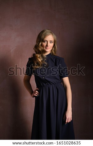 Vertical image of smiling woman pointing up and looking at camera while holding arm in pocket and posing near the wall in studio over gray background