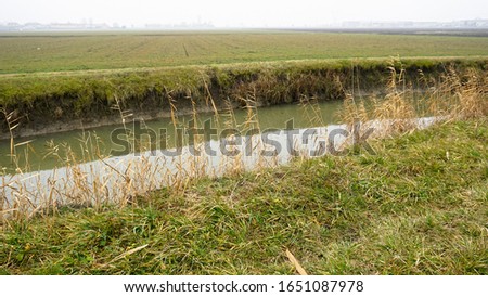 Beautiful view of a small countryside irrigation canal in Italy during an amazing dramatic Winter day. Grey sky, fog and green grass a bit frozen. Fields are resting waiting for the Spring