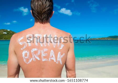 Young man with Spring Break message written in sunscreen on his back looking out over tropical island horizon