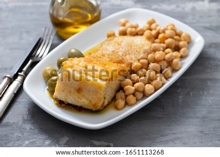 fried cod fish with chick pea and olives on white dish on ceramic background