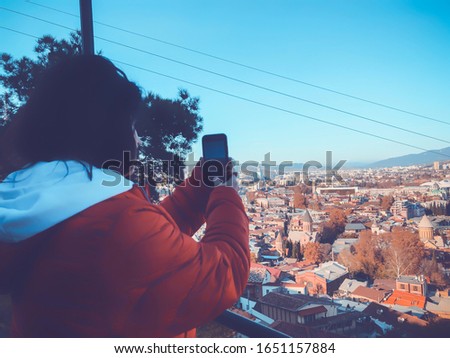 Girl in a red jacket taking pictures of the city. Beautiful landscape view of the old district with modern area. Old Tbilisi, winter in the city.