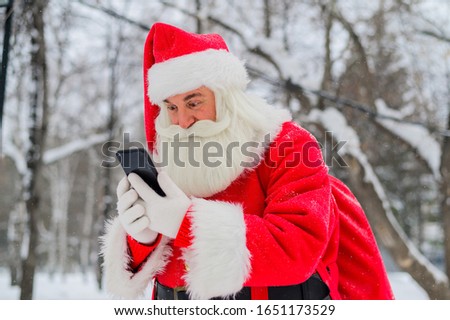 Surprised santa claus is using mobile outside. Santa Claus stands in the background of buildings in winter and calls on a smartphone. Snow in December.