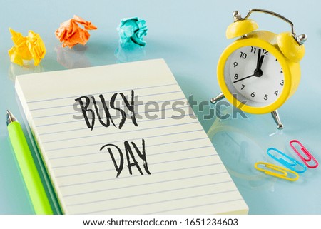 yellow alarm clock next to the notebook with the words "busy day" colored office staples, sticky notes, crumpled balls of paper.