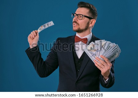 Man in black classic suit, red bow-tie, glases is holding some dollar bills, posing on gray studio background. Gambling, poker, casino. Close-up.