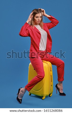 Blonde woman in white blouse, red pantsuit, high black heels. She touching hair, sitting on yellow suitcase, posing on blue background. Full length