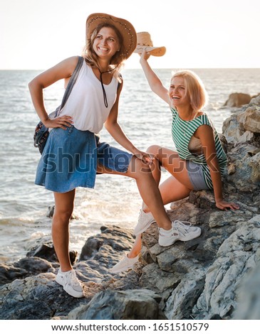 Two young cheerful women in hipsters hats on a rock on the coast of the sea. Summer landscape with girl, sea, Islands and orange sunlight. Travel and tourism with a friend. Twin sister, family