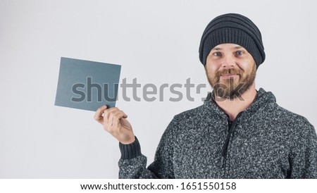 A Bearded Smiling Hipster Man On A White Background In A Knitted Hat Holds In His Hands A Blank Gray Carton Sign For Advertising. Copy space. High Quality Photo