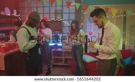 Mixed race attractive young urban people dancing together and using smartphones at local night party. Social media red heart like icons flowing around. Internet communication.