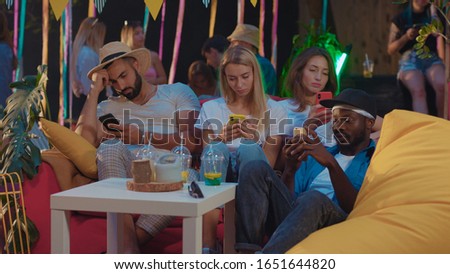 Group of bored young multiethnic friends using smartphones socializing online getting tired at the party celebration. Future communication. Mobile addiction.