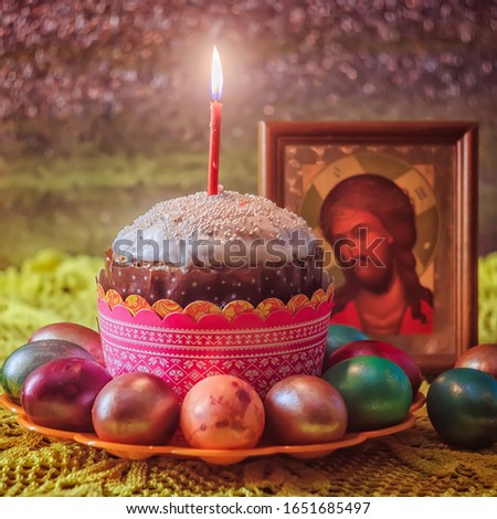 Butter cake with white icing and a burning red candle, colored painted eggs in a stand on the background of the icon of Jesus Christ. Concept: Orthodox Easter still life,traditional dishes