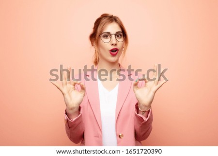 young pretty blonde woman feeling shocked, amazed and surprised, showing approval making okay sign with both hands. business concept