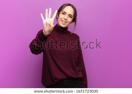young pretty woman smiling and looking friendly, showing number four or fourth with hand forward, counting down against purple wall