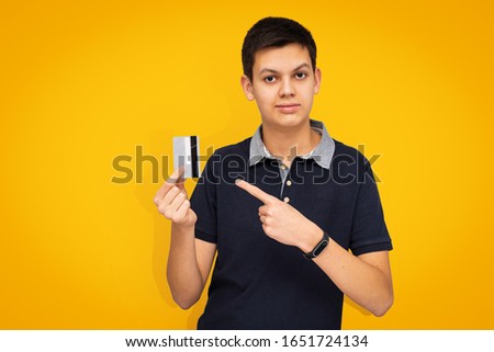 Portrait of attractive teenage boy pointing finger at credit card in his hand on yellow background. Cute optimistic teenager dressed in dark blue polo shirt presents and recommends use of bank cards