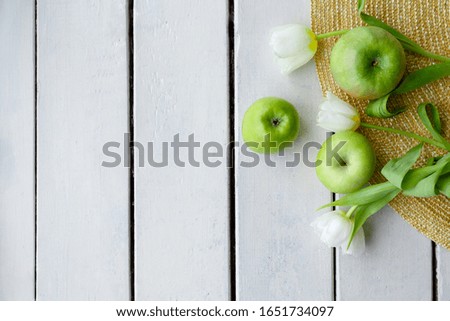green apples and white tulips lie on a straw hat and on a white background from wooden boards