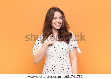 young pretty woman looking proud, confident and happy, smiling and pointing to self or making number one sign against orange wall