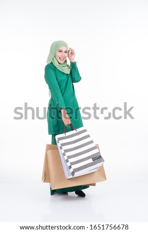 A beautiful Asian female model in a modern kurung carrying shopping bags isolated on white background. Eidul fitri festive preparation shopping concept. Full length portrait.