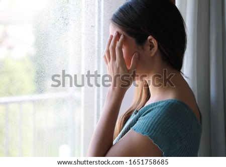 COVID-19 Pandemic Coronavirus Woman home isolation. Seasonal affective disorder concept. Sad depressed girl crying near the window at home in a rainy day, focus on model hand, indoor picture.