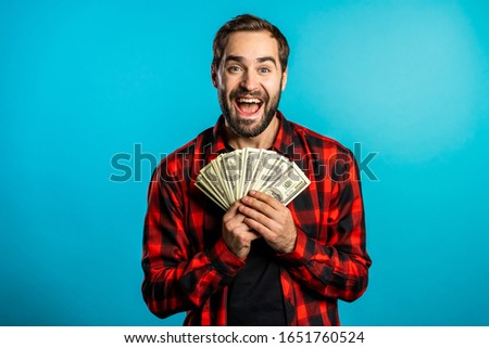 Satisfied happy excited man showing money - U.S. currency dollars banknotes on blue wall. Symbol of success, gain, victory.