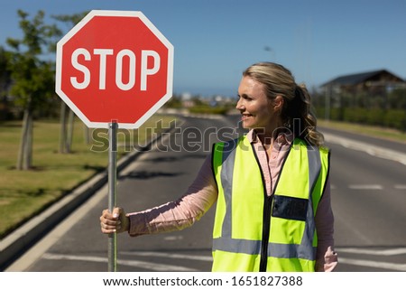 Front view close up of a blonde Caucasian woman wearing a high visibility vest and holding a stop sign, standing in the road and looking to the side on a pedestrian crossing, waiting to help children